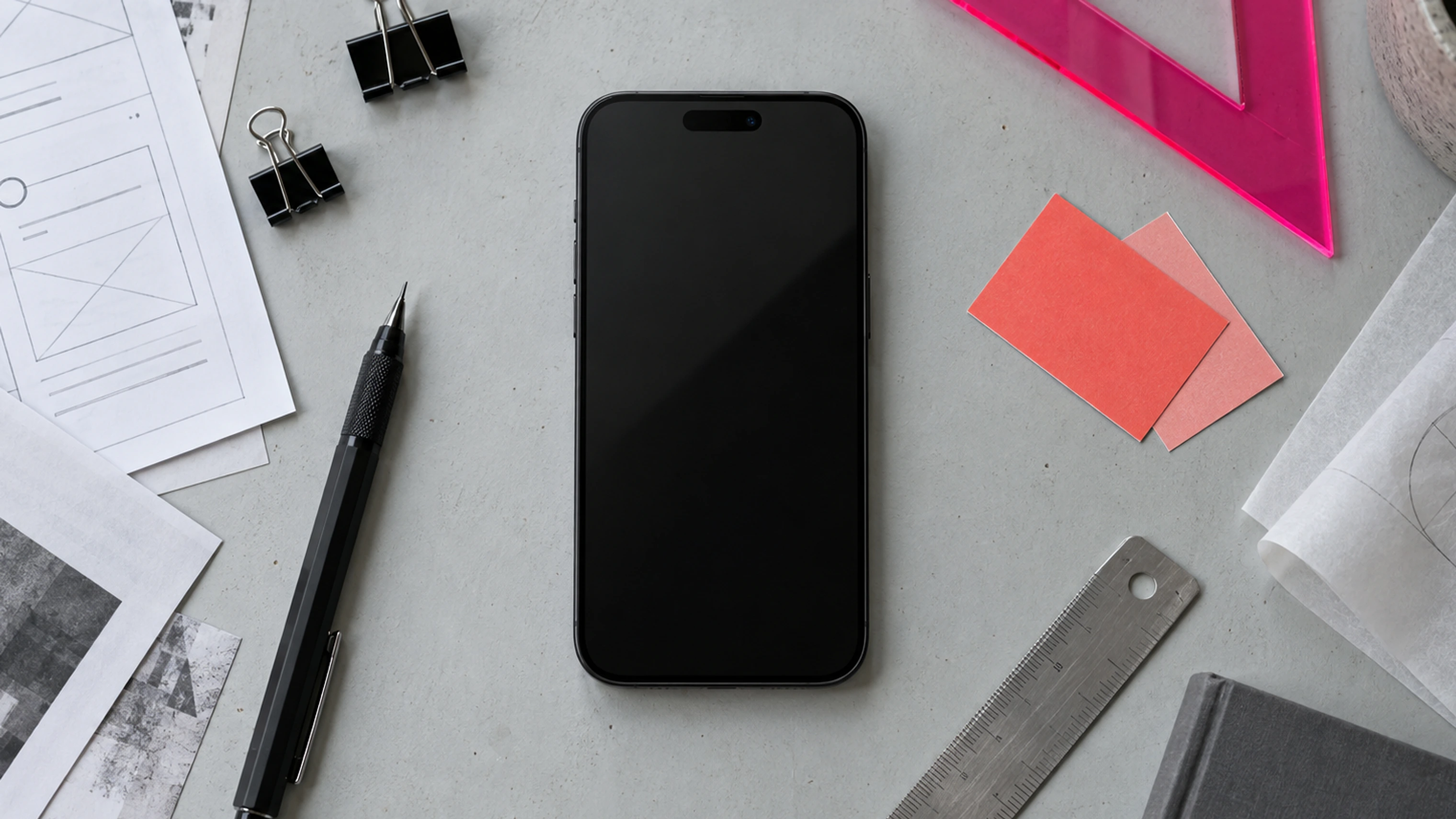 An iPhone on a design desk surrounded by sketches, tools, coral swatches and a vivid pink set square.